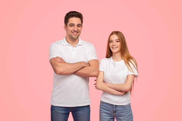 Self assured young couple in similar clothes folding arms and looking at camera with smile while standing against pink background together - Foto, Bild