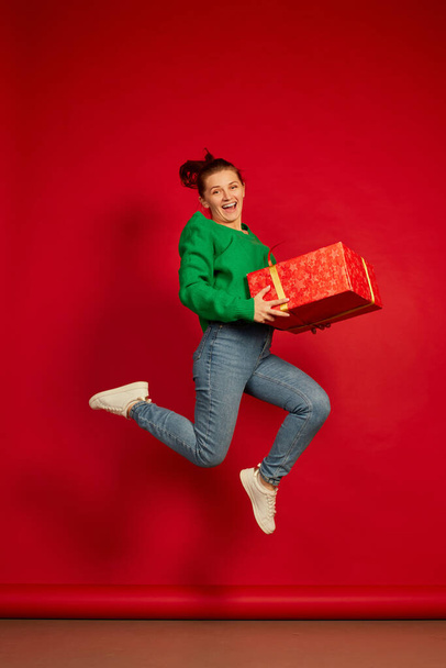 Portrait of happy smiling woman in green sweater jumping in joy with present box isolated over red studio background. Concept of holidays, happiness, emotions, facial expression, joy, celebration - Φωτογραφία, εικόνα