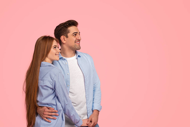 Positive young loving couple in similar casual clothes embracing smiling and looking away against pink background - Photo, image