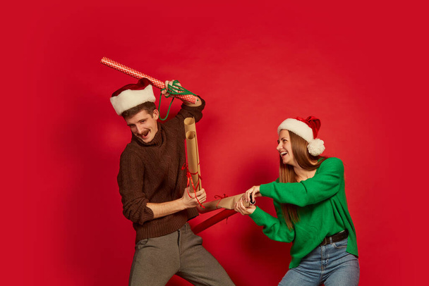 Portrait of cheerful young people, man and woman fighting in joy with wrapping paper isolated over red background. Concept of holidays, happiness, emotions, facial expression, joy, celebration - Фото, изображение
