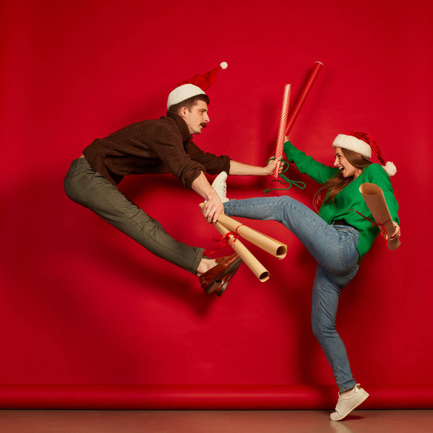 Portrait of cheerful young people, man and woman jumping with wrapping presents paper isolated over red background. Concept of holidays, happiness, emotions, facial expression, joy, celebration - Fotografie, Obrázek