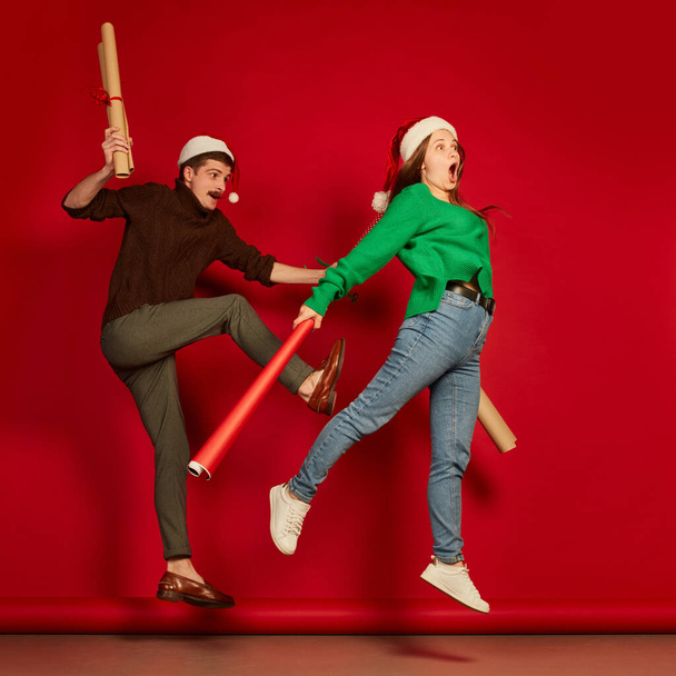 Portrait of cheerful young people, man and woman with wrapping presents paper isolated on red background. Funny emotions . Concept of holidays, happiness, emotions, facial expression, joy, celebration - Foto, Imagen