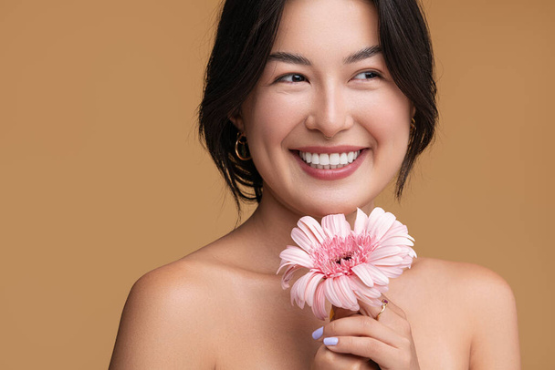 Happy young Asian woman with bare shoulders holding fresh pink gerbera flower and looking away with toothy smile against brown background - Foto, imagen