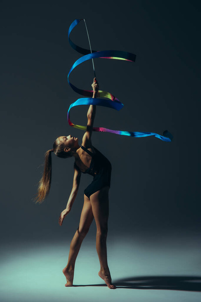 Jimnastikçi kız siyah elbiseli ışıkla aydınlatılıyor. Elinde kurdeleyle jimnastik yapıyor.. - Fotoğraf, Görsel