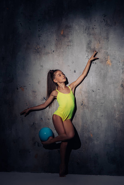 Gymnast child girl lit by light in yellow bodysuit performs gymnastic exercises with ball on dark background. - Photo, Image