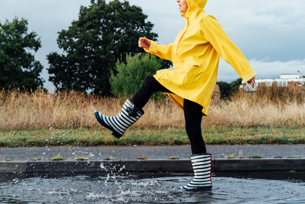 Woman having fun on the street after the rain. Cropped woman wearing rain rubber boots and yellow raincoat walking into puddle with water splash and drops. Fall weather. Selective focus. - Foto, afbeelding