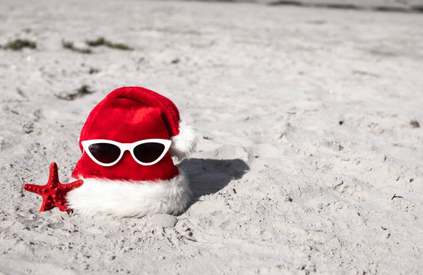 Kerstachtergrond Kerstmuts op het strand met zeesterren en zonnebrillen.Kerstkaart en adventskalender concept. Reisticket verkoop concept voor kerstvakantie kopieerruimte.Hoge kwaliteit - Foto, afbeelding