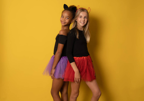 Εικόνα των γελαστών πολυεθνικών κοριτσιών με μαύρα και πολύχρωμα αποκριάτικα κοστούμια που απομονώνονται πάνω από κίτρινο τοίχο - Φωτογραφία, εικόνα