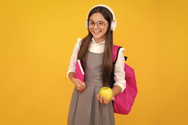Iskolásgyerek, tinédzser lány táska tartsa alma és könyv elszigetelt sárga. Boldog arc, pozitív és mosolygós érzelmek tinédzser lány - Fotó, kép