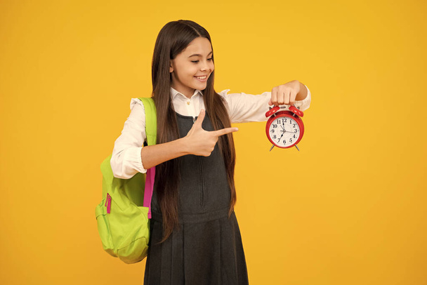 Zurück zur Schule. Teenager-Schulmädchen mit Rucksack halten Wecker, Zeit zum Lernen. Schulmädchen auf gelbem Hintergrund. Glückliches Mädchengesicht, positive und lächelnde Emotionen - Foto, Bild