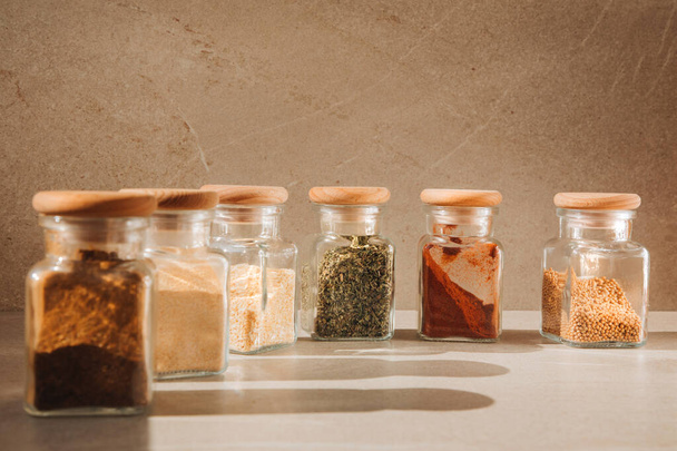 A group of seasonings in glass jars on a light stone background with shadows. Paprika, herbs, mustard, garlic, front view, selective focus - Valokuva, kuva