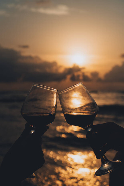 Close-up silhouette of two glasses of wine clinking against the background of a beautiful sunset on the sea. Concept of celebration, romance, friendship. Vertical photo - Foto, immagini