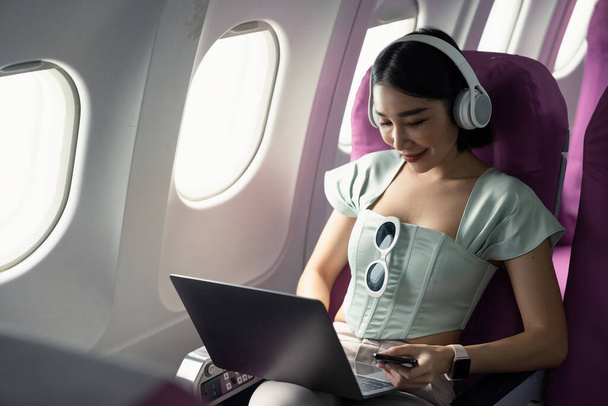 Avion passager asiatique femme d'affaires travaillant en cabine d'avion pendant le vol avec ordinateur portable wifi écouter de la musique avec écouteurs. - Photo, image