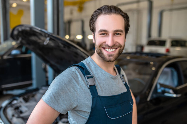 Portrait of a cheerful automotive service mechanic posing for the camera among the cars in the workshop - Fotoğraf, Görsel