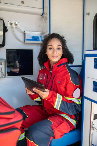 Pleased healthcare worker with the tablet computer sitting in the medical emergency vehicle and looking before her - Valokuva, kuva