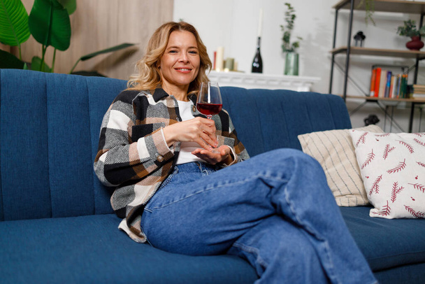 Portrait d'une jolie femme blonde d'âge moyen buvant du vin assis sur un canapé à la maison - Photo, image