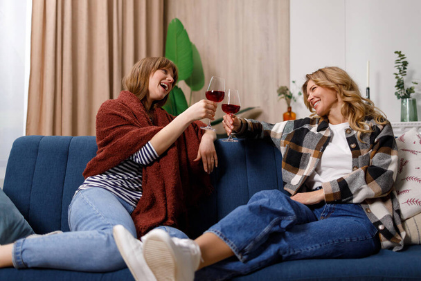 Mature woman and her daughter celebrating something and cheers glasses of wine with each other sitting on the sofa in the living room - Fotoğraf, Görsel