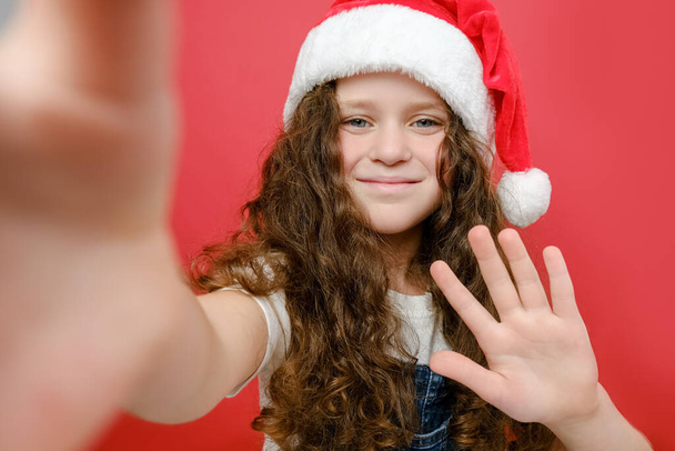 Portrait of smiling little girl child looks surprisingly feels glad makes selfie, wears Santa Claus hat, posing isolated over red color background wall in studio. Happy New Year 2023 holiday concept - Foto, immagini