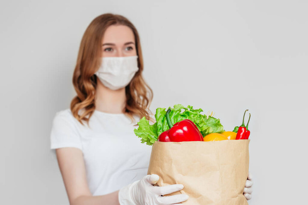 Young woman courier wearing a medical mask holds a paper bag with food, vegetables, pepper, chilli, lettuce isolated over white, light gray background, home delivery, quarantine, food delivery concept - Φωτογραφία, εικόνα