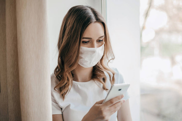 A young woman sits on a windowsill, looks at a mobile phone and reads a message, searches for online remote work, coronavirus, quarantine, isolation, safe work, communication - Photo, image