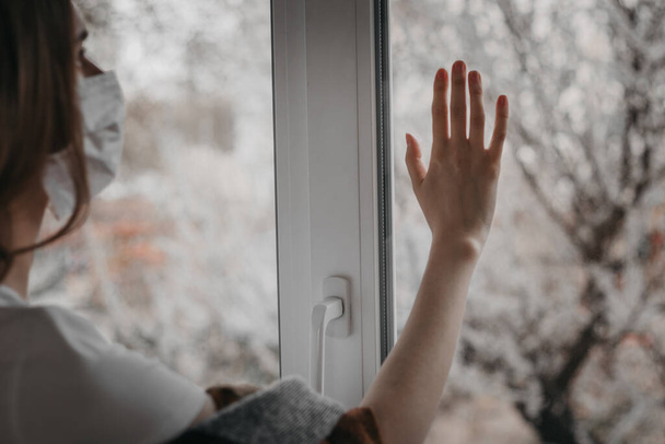 Νεαρή γυναίκα με ιατρική μάσκα κοιτάζει έξω από το παράθυρο και κρατά το χέρι της στο τζάμι του παραθύρου, καραντίνα, απομόνωση, κορωναϊός, να είστε ασφαλείς. - Φωτογραφία, εικόνα