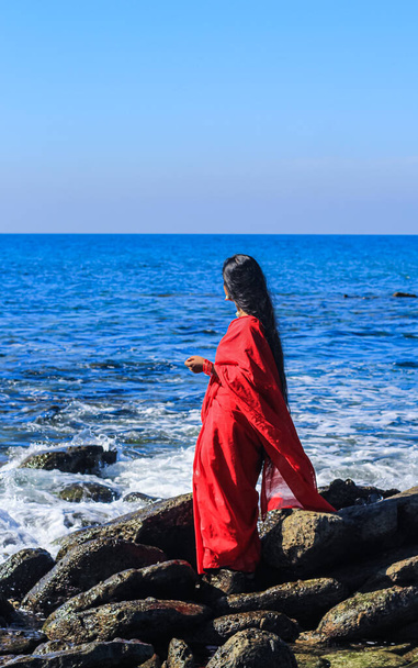 Молодые женщины в красном сари на пляже. Девушка в традиционном индийском сари среди скал и наслаждается свободой и закатом или восходом солнца. Красивая индийская женщина на пляже в сари. - Фото, изображение