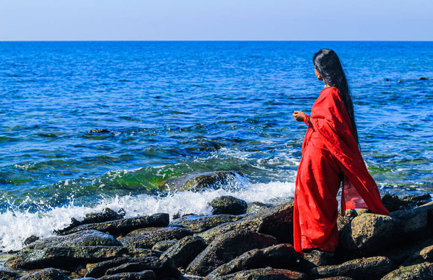 Young women wearing a red saree on the beach. Girl in traditional Indian sari among the rocks and enjoying the freedom and the sunset or sunrise. Beautiful Indian woman on the beach wearing a sari. - Foto, Imagen