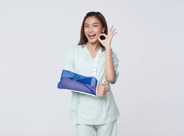 Asian teenage woman wearing a patient gown put on a cast due to injury from an accident showing hand okay with an impressive smile.insurance concept. - Photo, Image