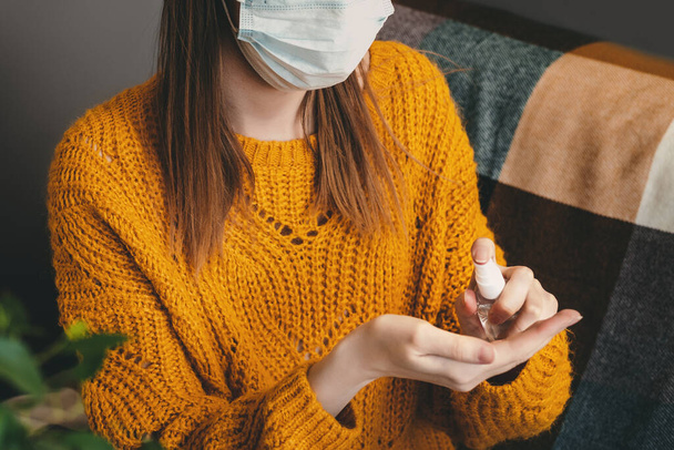 A girl in a medical mask sits on a sofa at home in isolation and uses antiseptic hand gel, hygiene with coronovirus. A young woman sits at home in quarantine and washes her hands with an alcohol spray - Photo, Image