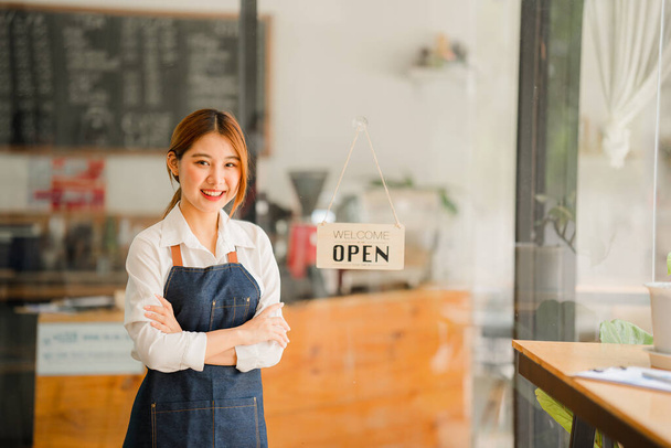 A younger shopkeeper standing in front of her shop. Open a shop. A beautiful Asian woman in an apron holds a tablet and stands in front of a cafe door with an open sign. business owner startup ideas - Photo, Image