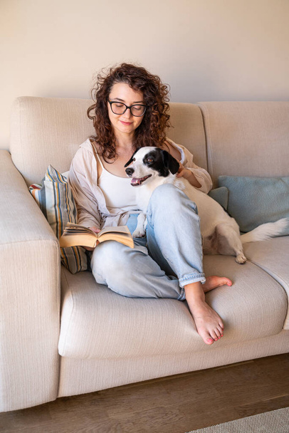 Beautiful curly girl with glasses sitting on sofa with book and her black and white adopted dog. Light sunny living room. reading with smiling pet. Happy relaxed weekend moment at home with dog - Фото, изображение