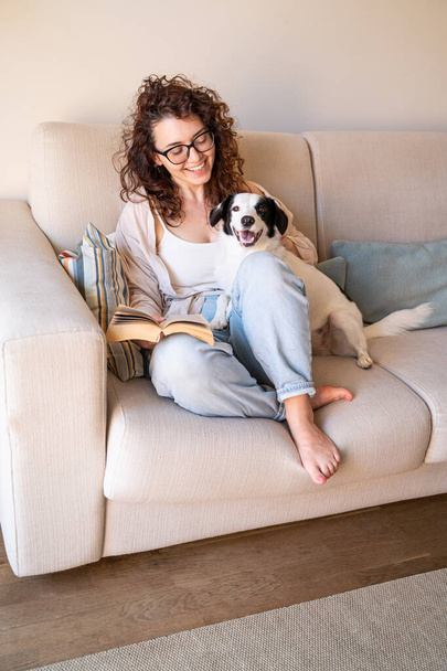 Beautiful curly girl in glasses sitting on sofa in living room with her young black and white dog reading a book. Smiling looking at happy dog. Relaxed weekend time with pet. blue jeans, casual shirt - Photo, Image