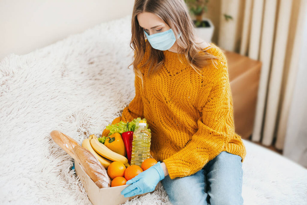 Jong meisje vrijwilliger in een medisch masker houdt een kartonnen doos met veganistisch voedsel, groenten en fruit en zit op een bank, levering aan huis, coronovirus, quarantaine, blijf thuis concept - Foto, afbeelding