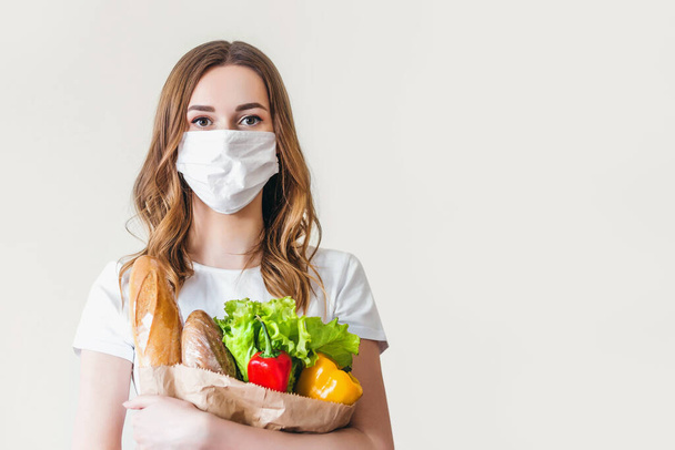 Young woman in medical mask holds a paper bag with food, fruits and vegetables, pepper, baguette, lettuce over grey background, home delivery, coronovirus, quarantine, stay home concept, copy space - Fotoğraf, Görsel