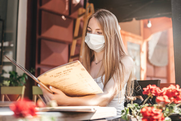 Portret van een blank blond meisje met een beschermend medisch masker zittend aan een tafel in een café op straat, het lezen van een menu en het maken van een maaltijd orde, quarantaine regels voor de coronavirus epidemie - Foto, afbeelding