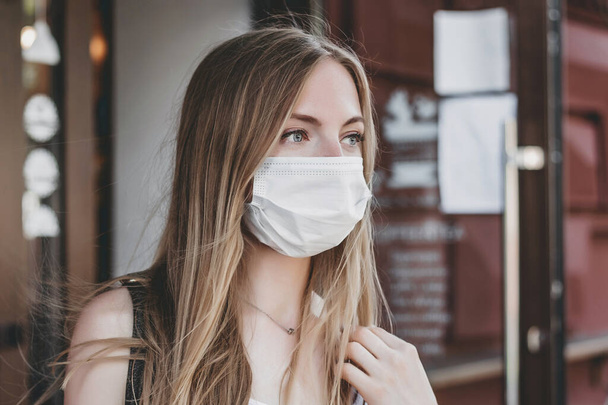 Közelkép egy fehér szőke lányról, aki orvosi maszkot visel, és a város utcáin áll egy kávézó homlokzatán. - Fotó, kép