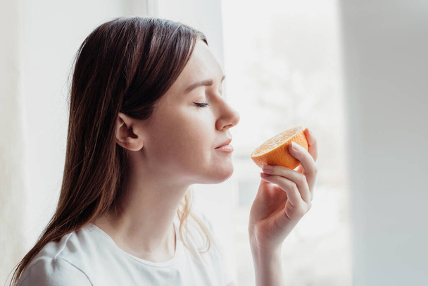 Verlies van geur concept. Blanke jonge vrouw staat bij het raam en ruikt een sinaasappel - Foto, afbeelding
