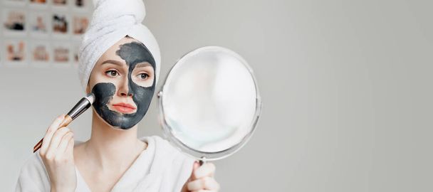 A young girl in a white bathrobe with a towel on her head holds a mirror in her hands and applies a natural black mask with activated carbon for problem skin, quarantine isolation, home spa treatments. banner, copy space - Photo, image