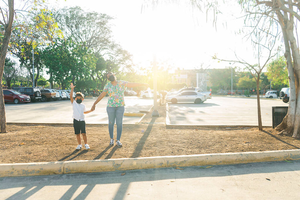 Latin mother and son with masks walking outdoors in a park in Managua, Nicaragua, during the sunset - Foto, Imagen
