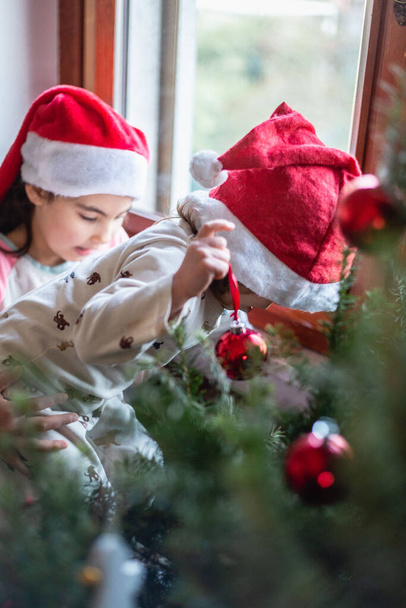 Petite sœur de 2 ans et sœur aînée de 8 ans décorant l'arbre de Noël à la maison pendant la journée avec des décorations rouges - Photo, image
