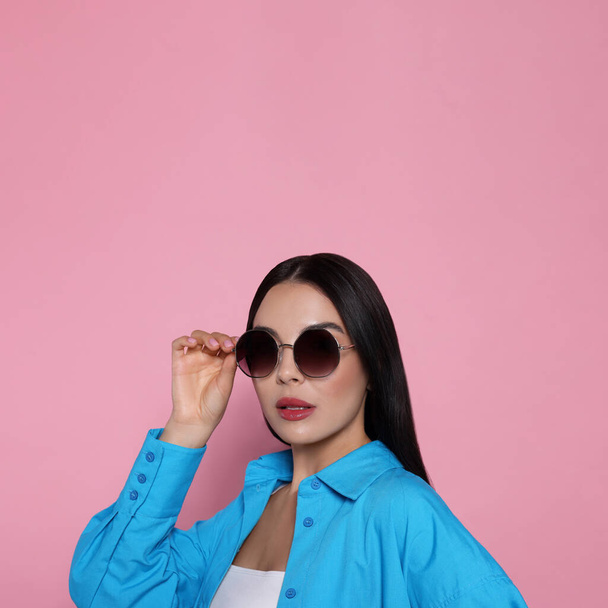 Attractive serious woman touching fashionable sunglasses against pink background. Space for text - Photo, image