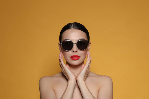 Attractive woman in fashionable sunglasses touching face against orange background - Фото, изображение