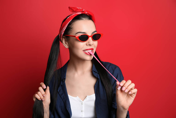 Fashionable young woman in pin up outfit chewing bubblegum on red background - Foto, afbeelding