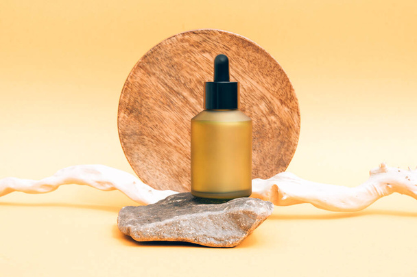 Matte glass dropper bottle with organic cosmetic oil or face serum against light yellow background with stone and wooden stick as decorations. Natural skin care products. Mockup - 写真・画像
