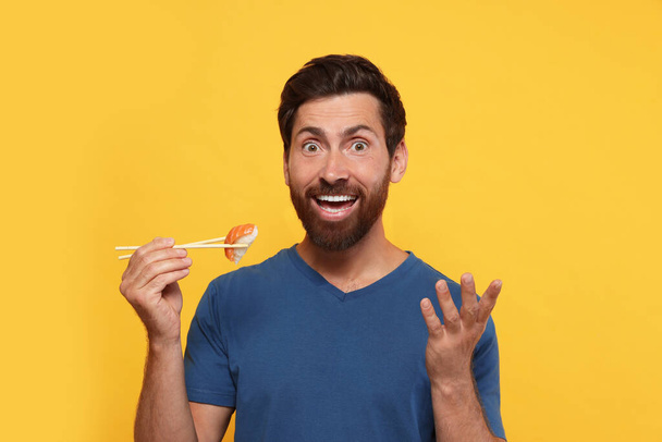 Emotional man holding sushi with chopsticks on orange background - 写真・画像
