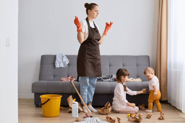 Indoor shot of tired female with bun hairstyle washing floor with mop at home, children making mess in living room, exhausted mother trying calm down and relax. - Foto, Imagen