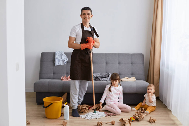 Full length portrait of smiling positive Caucasian man wearing brown apron washing floor with mop at home, his kids playing in living room in mess, guy looking at camera with happiness. - Fotoğraf, Görsel