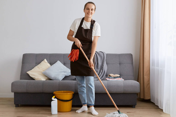 Full length portrait of smiling attractive woman wearing jeans and brown apron, washing floor with mop at home, looking at camera with happy positive emotions, likes to do work about the house. - Foto, Imagem