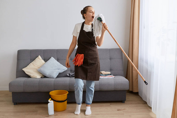 Indoor shot of attractive woman with bun hairstyle washing floor with mop at home, having fun while cleaning apartment, using mop as a microphone, singing while tidy up. - Photo, image