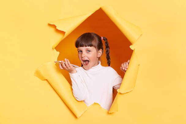 Indoor shot of excited amazed little girl with pigtail breaking through yellow paper and holding cell phone, recording voice message, expressing positive emotions. - Фото, зображення